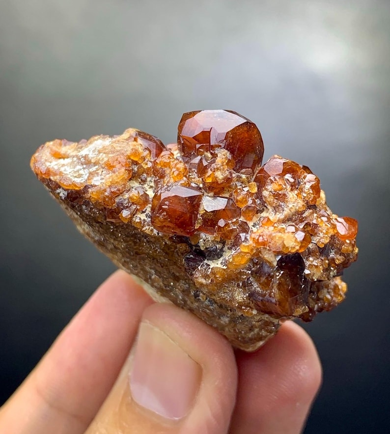 May include: Close-up of a cluster of orange-brown garnet crystals. The crystals have a glossy, faceted appearance and are set on a rough, brown matrix. The mineral specimen is held in a hand.