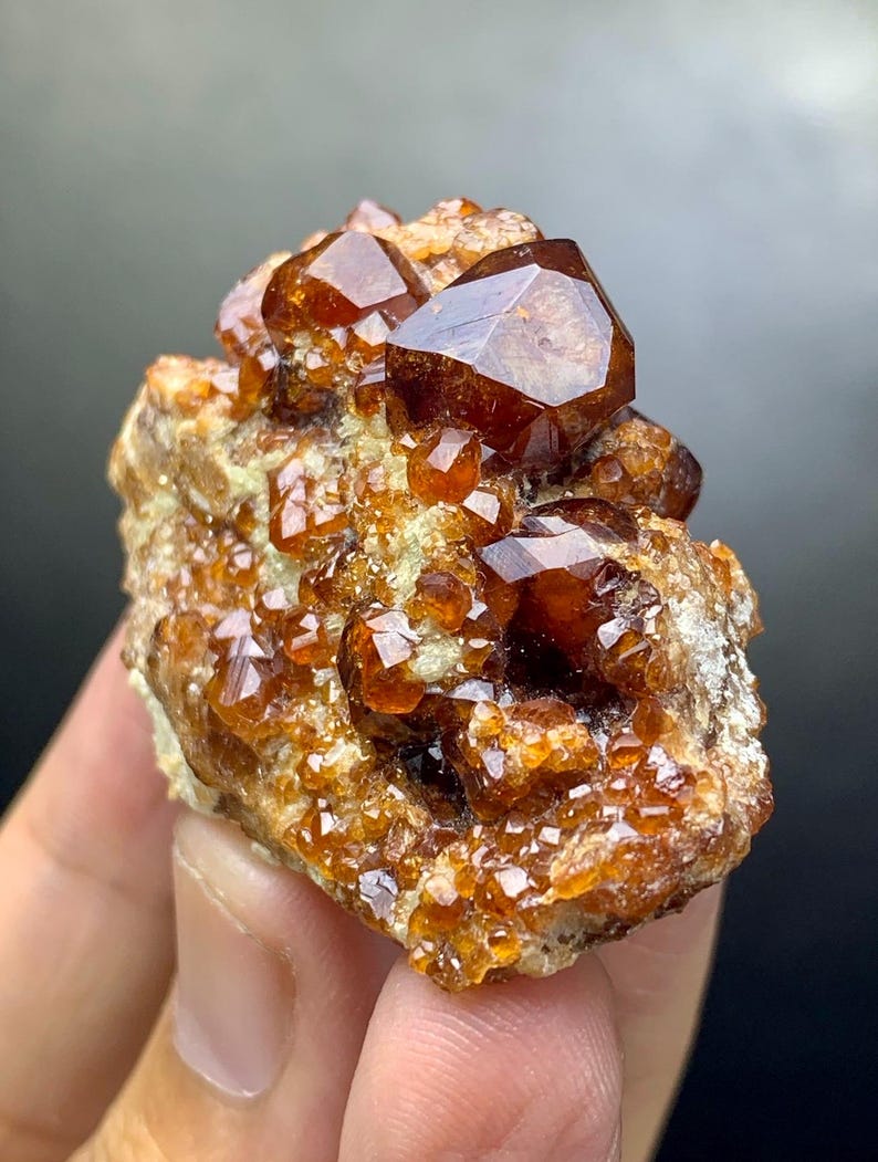 May include: A cluster of lustrous, reddish-brown garnet crystals. The crystals have a geometric shape and are set against a rough, beige matrix. The gemstone cluster is held in a hand, with a dark background.