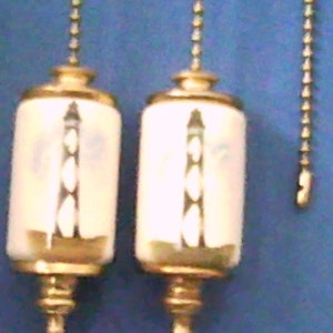 May include: Two white ceramic light pull chains with gold accents and a lighthouse design. The chains are attached to gold metal rings.