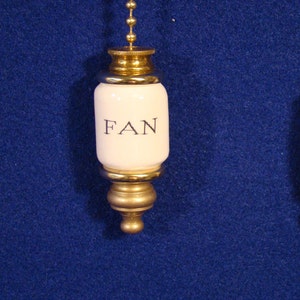 May include: A white ceramic fan pull with a gold chain and a gold metal base. The pull has the word "FAN" printed on it in black.