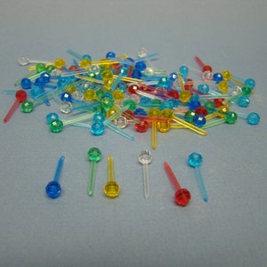 May include: A selection of colourful plastic cocktail picks with faceted ball tops. The picks are in various colours, including red, blue, green, yellow, and clear. Some picks are scattered, while others are arranged in a row. The background is a light blue.