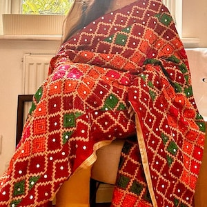 Op de afbeelding: Een levendige rode sjaal met een geometrisch patroon in oranje, groen en wit. De stof is versierd met kleine, reflecterende versieringen. De sjaal heeft een gouden rand langs de randen. Dit textiel is geschikt voor speciale gelegenheden.