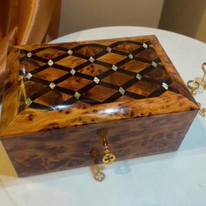 May include: A wooden jewellery box with a geometric inlaid design on the lid. The box is a warm brown colour with a keyhole and a decorative key. The lid features a pattern of dark brown diamonds and small, light-coloured squares.
