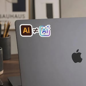 May include: A rectangular sticker applied to a grey laptop lid. The sticker features a brown square icon with the text Ai, a white not-equal sign, and a colourful iridescent square icon with the text Ai and three small white star symbols above it.