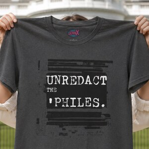 May include: A charcoal gray short-sleeved t-shirt featuring a black rectangular graphic with the white text UNREDACT THE PHILES. The design includes horizontal black redaction lines above and below the text. The inner neck label reads JENNX.