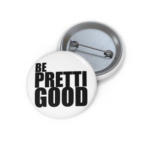 May include: Two white round buttons with a pin back. One button is in focus, displaying the phrase "BE PRETTI GOOD" in bold black letters. The other button is slightly out of focus, showing the pin back.