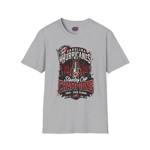 May include: A heather gray short-sleeve t-shirt featuring a graphic of the Stanley Cup with the text Carolina Hurricanes Stanley Cup Champions 2025-2026 Season. The design includes red and black accents with the team logo and years 2006 and 2025.