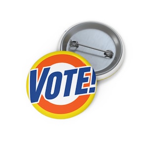 May include: A round pin-back button featuring the word VOTE! in bold blue letters centered on a white circle, surrounded by an orange ring and a yellow outer border. The back of the button is silver-toned metal with a horizontal pin mechanism.