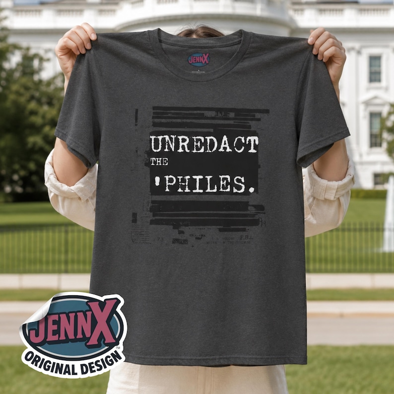 May include: A charcoal gray short-sleeved t-shirt featuring a black rectangular graphic with the white text UNREDACT THE PHILES. The design includes horizontal black redaction lines above and below the text. The inner neck label reads JENNX.