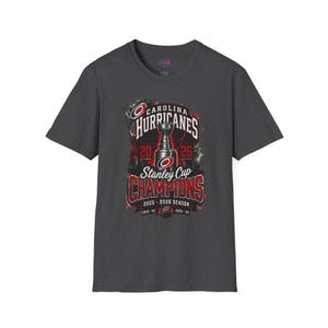 May include: A dark gray short-sleeve t-shirt featuring a graphic of the Stanley Cup with the text Carolina Hurricanes Stanley Cup Champions 2025-2026 Season. The design includes red and white accents, a hockey puck, and the team logo.