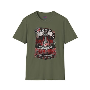 May include: An olive green short-sleeve t-shirt featuring a graphic print of the Carolina Hurricanes Stanley Cup Champions 2025-2026 season. The design includes the team logo, a trophy illustration, and the text 2025-2026 season and 2005-06.
