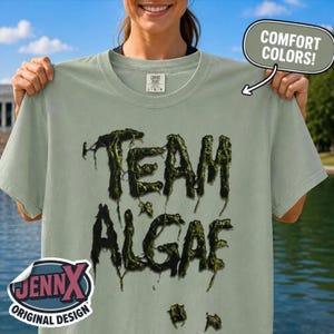 May include: A sage green short-sleeve t-shirt featuring the text TEAM ALGAE in a dark, dripping, textured green font. The shirt has a Comfort Colors label at the neck and a JennX Original Design logo in the corner. The fabric is a solid, muted tone.