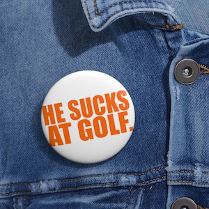 May include: A white button with the orange text "HE SUCKS AT GOLF." The button is pinned to a blue denim jacket. The button is round and has a glossy finish.