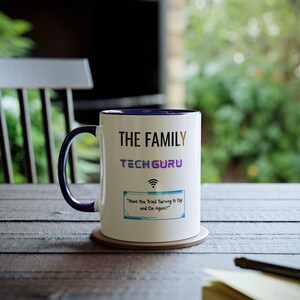 Puede incluir: Taza de cerámica blanca con asa y borde azules. La taza presenta el texto "THE FAMILY TECH GURU" en negro y morado, junto con un símbolo Wi-Fi y la frase "Have You Tried Turning It Off and On Again?"