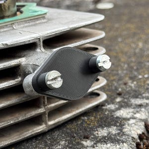 Könnte beinhalten: Nahaufnahme einer silbernen Metallmotorkomponente mit einer schwarzen Abdeckung, die mit zwei silbernen Schrauben befestigt ist. Die Komponente hat mehrere horizontale Rippen und eine grüne Dichtung.