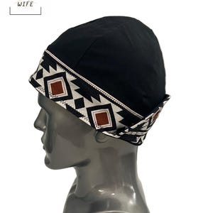 Puede incluir: Gorra de soldador negra con un patrón geométrico en blanco, marrón y negro. La gorra tiene un borde doblado y presenta el texto "THE WELDER'S WIFE" en blanco.