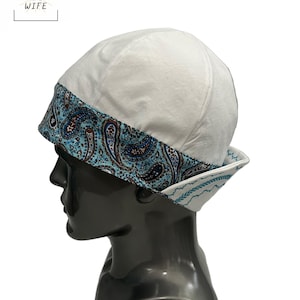 May include: White welding cap with a blue and brown paisley patterned brim. The cap is displayed on a black mannequin head. The text "THE WELDER'S WIFE" is in the upper left corner.