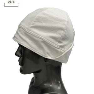 May include: A white welding cap is displayed on a black mannequin head. The cap is made of a soft fabric and has a folded brim. The text "THE WELDER'S WIFE" is in a white box in the upper left corner.