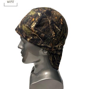 May include: A camouflage welding cap with a forest pattern of brown, green, and orange. The cap is on a mannequin head. The text "THE WELDER'S WIFE" is in a white box in the upper left corner.