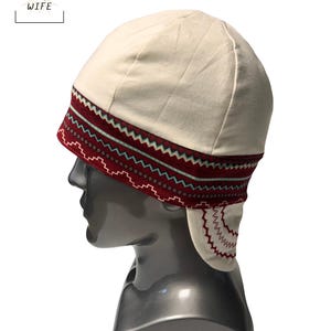 May include: Cream-colored welding cap with a decorative band featuring a zigzag pattern in burgundy, teal, and white. The cap has a rounded crown and a flap that extends to cover the ears. The text "THE WELDER'S WIFE" is in the upper left corner.