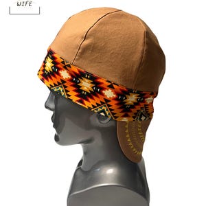 Puede incluir: Gorra de soldador marrón con una banda estampada con diseños geométricos naranjas, amarillos, negros y blancos. La gorra tiene una corona redondeada y orejeras. El texto "THE WELDER'S WIFE" está en la esquina superior izquierda.