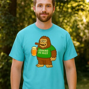 May include: A turquoise t-shirt featuring a cartoon Bigfoot character holding a beer and a cigar. The Bigfoot wears a green shirt with the text "LOOKING FO HUZZ". The Bigfoot is brown with tan fur and a yellow face.