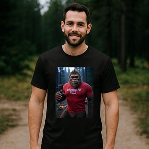 May include: Black t-shirt featuring a graphic of a Bigfoot-like creature holding a bottle and cigar, wearing a red shirt with the text "LOOKING FO HUZZ". The image is set against a forest backdrop.