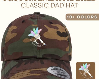 May include: Camouflage classic dad hat with a custom embroidered design. The hat features a colorful paintbrush logo and the text "Your Custom Design." The hat is adjustable and available in 10+ colors.