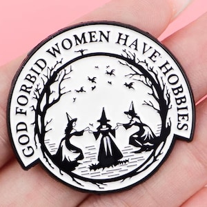 Pode incluir: Um alfinete redondo de esmalte preto e branco com o texto "GOD FORBID WOMEN HAVE HOBBIES". O alfinete apresenta três bruxas em círculo, rodeadas por árvores e pássaros. Um acessório divertido para o Halloween ou para estética de bruxas.