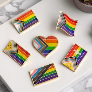Può includere: Una collezione di spille smaltate con bordi dorati raffiguranti vari disegni di bandiere Pride. Le spille includono bandiere arcobaleno, bandiere con motivi geometrici e una spilla a forma di cuore. Le spille sono esposte su una superficie bianca.
