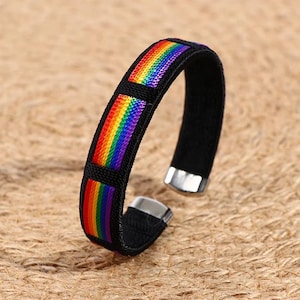 Puede incluir: Una pulsera negra con un diseño de rayas arcoíris. La pulsera presenta una banda negra con un colorido patrón de arcoíris en rojo, naranja, amarillo, verde, azul y morado. La pulsera tiene detalles metálicos plateados.