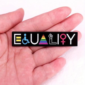 Può includere: Una spilla rettangolare nera con la scritta "EQUALITY" in lettere bianche. La spilla presenta simboli colorati tra cui un'icona di sedia a rotelle, un arcobaleno, un pugno alzato e un simbolo femminile. La spilla è tenuta in una mano.