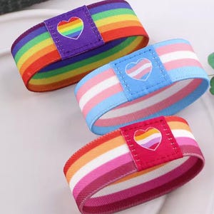 Puede incluir: Tres pulseras elásticas coloridas. Una presenta un diseño de arcoíris con un corazón, otra tiene los colores de la bandera transgénero con un corazón, y la tercera tiene los colores de la bandera lesbiana con un corazón. Las pulseras están sobre una superficie blanca.