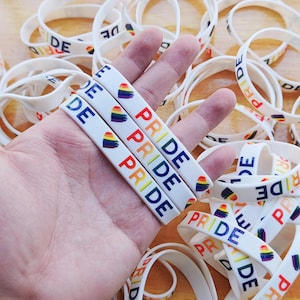 Op de afbeelding: Witte rubberen polsbandjes met het woord "PRIDE" in regenboogkleuren en een regenboog hart ontwerp. Andere polsbandjes op de achtergrond hebben de woorden "PRIDE" en "DE" erop gedrukt. De polsbandjes zijn een symbool van LGBTQ+ trots.