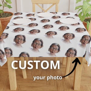 May include: A white tablecloth featuring a repeating pattern of a woman's face wearing glasses. The text "CUSTOM your photo" is displayed in large white letters. The tablecloth is on a wooden table with two white plates.