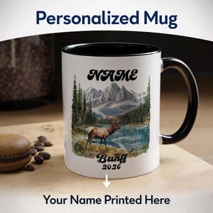 May include: A personalized white mug with a black interior and handle. The mug features a scenic illustration of mountains, a lake, and an elk. The text "NAME" and "Banff 2026" are printed on the mug, with the words "Your Name Printed Here" below.