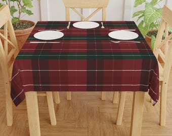 May include: A rectangular tablecloth with a red, green, and white plaid pattern. The tablecloth is draped over a wooden table set with white plates and silverware. The table is surrounded by wooden chairs.