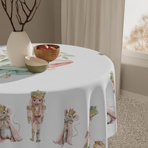 May include: A white round tablecloth featuring watercolor illustrations of a nutcracker and mice in regal attire. The design includes a vase, bowls, and a basket of apples. The tablecloth is suitable for a dining table.