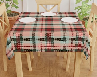May include: A plaid tablecloth in shades of red, green, and pink covers a wooden table. Three white plates are set on the table. The tablecloth has a classic plaid pattern, with intersecting lines creating a grid-like design. The table is surrounded by wooden chairs.