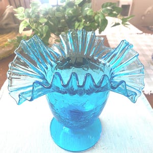 May include: A vibrant blue glass vase with a ruffled rim. The vase has a crackle finish and sits on a round base. The ruffled rim adds a decorative touch to this vintage-style vase.