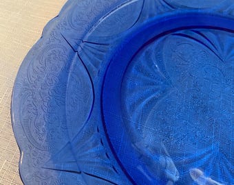 May include: Close-up of a vibrant blue glass plate with a scalloped edge. The plate features an embossed floral pattern around the rim and a smooth, raised center. The plate is likely vintage and suitable for serving.