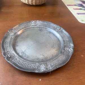 May include: A silver-colored, round plate with an ornate, embossed border. The plate has a slightly concave center and is likely made of metal. The decorative border features floral and geometric patterns.
