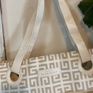 May include: A Givenchy tote bag with a geometric pattern in beige and white. The bag features white leather handles and a small rectangular label that reads "Givenchy Parfums". The bag has a circular handle attachment.