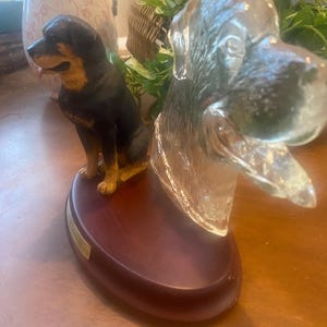 May include: A decorative Rottweiler dog figurine with a black and tan coat, sitting on a wooden base. Beside it is a glass dog head sculpture. The base has a gold nameplate.