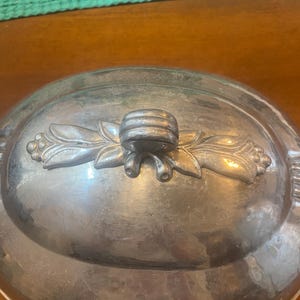 May include: A silver-colored, oval serving dish lid. The lid features a decorative floral design with a central handle. The dish appears to be made of metal and has a slightly aged appearance, suggesting it may be vintage.