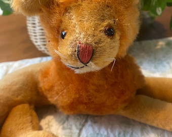 May include: A vintage orange plush lion toy with a red nose and black eyes. The lion has a fluffy mane and is lying down on a patterned surface. The toy is made of soft, worn material.