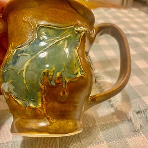 May include: Handmade ceramic mug with a unique leaf design. The mug features a brown exterior with a green and gold leaf pattern. The handle is curved and the mug has a glossy finish. A perfect addition to any kitchen.