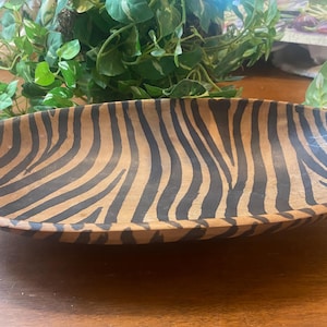 May include: Oval wooden serving bowl with a zebra stripe pattern in black and tan. The bowl is empty and sits on a wooden surface. The bowl is a decorative piece.