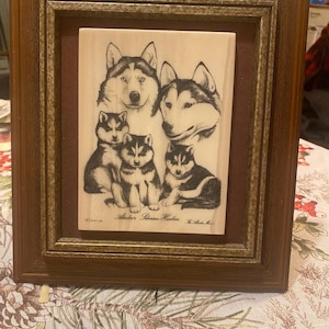 May include: A framed artwork featuring a family of Siberian Huskies. The artwork depicts two adult Huskies and three puppies in a black and white sketch style. The frame is a dark brown color.