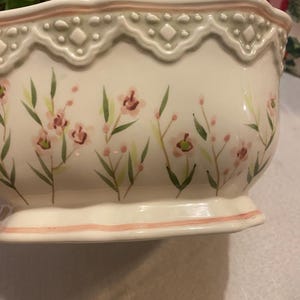 May include: A white ceramic bowl with a scalloped edge and floral design. The bowl features pink flowers and green leaves painted on the exterior. A pink stripe accents the base and rim.